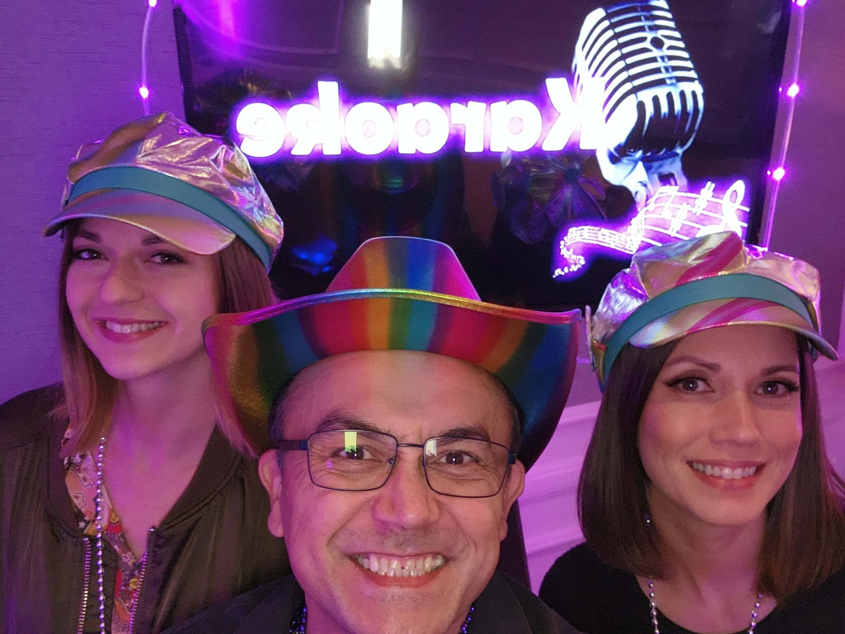 Liz Eryn Roman touch up jpeg RESZD - IG ratio Karaoke Dj Services team members. Three people wearing colorful hats smile in front of a neon "Karaoke" sign and microphone decoration, celebrating in a festive setting