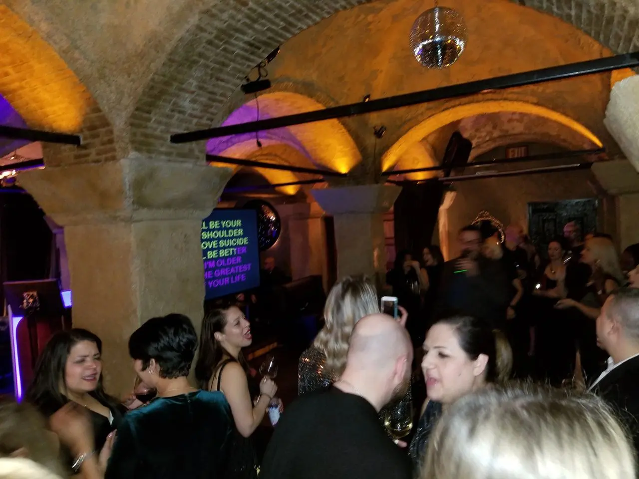 Mr Dj Services A group of people socializes at a dimly lit indoor party with arched ceilings and a disco ball; a karaoke screen displays song lyrics in the background.