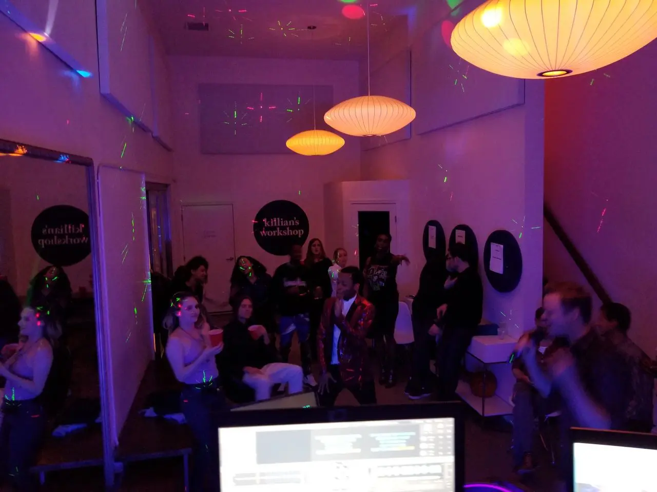 Mr Dj Services Karaoke dj A group of people socialize and dance in a dimly lit room with colorful lights and hanging lamps; computer monitors are visible in the foreground.