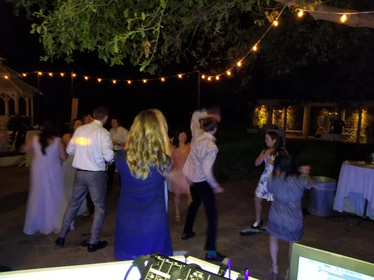 Mr Dj Services weddings A group of people dance outdoors at night under string lights, with trees and a table visible in the background.