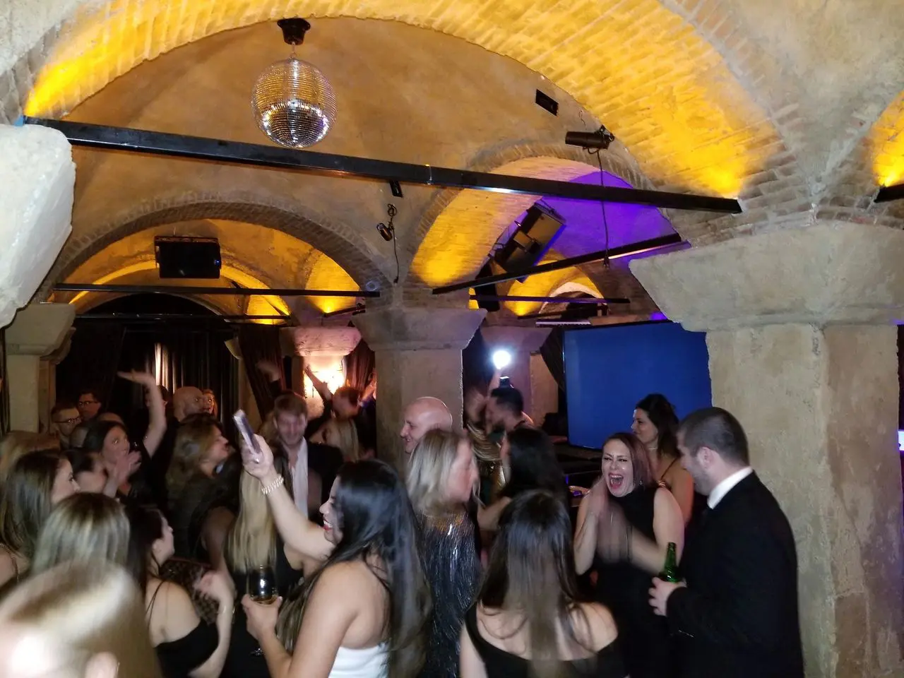 The Hollywood Roosevelt A group of people in formal attire dance and socialize under arched stone ceilings at a lively indoor party with yellow accent lighting.