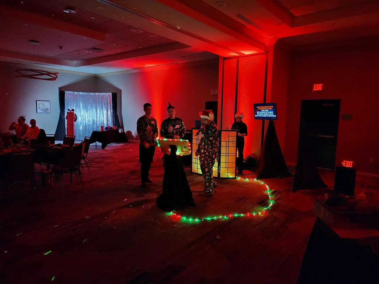 Holiday Party Christmas Four people in costumes stand near a lit DJ booth in a dimly lit room with red lighting and string lights on the floor; guests are seated at tables in the background.