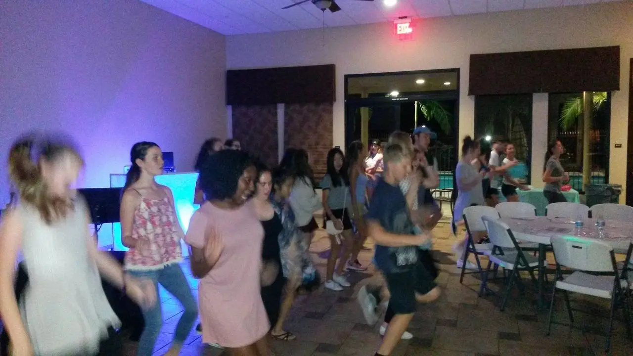 Karaoke Dj Party for kids A group of people dance together in a room with tables and chairs; some are blurred due to movement. The room is lit with blue and white lighting.