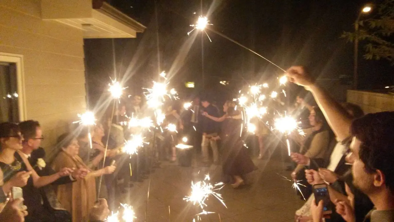 Wedding Dj Service People form two lines holding sparklers at night while a couple dances in the middle of a pathway outside a building.