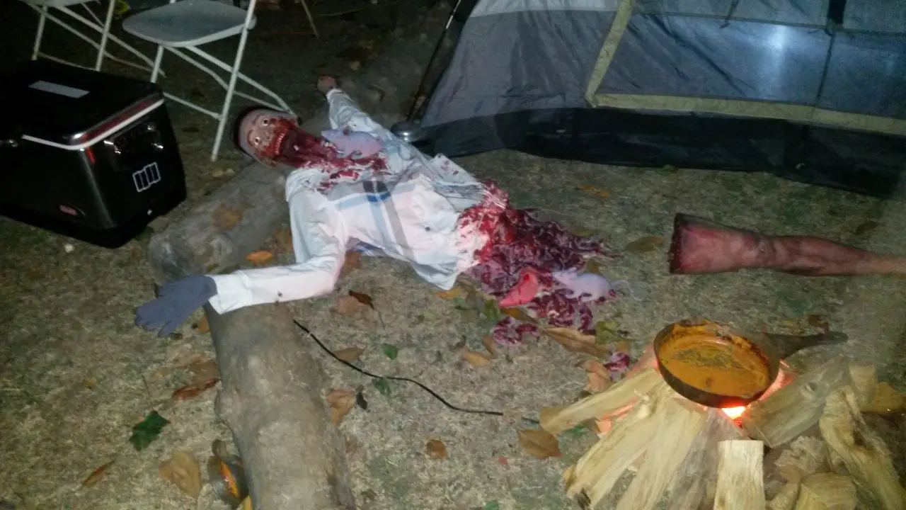 Halloween Dj Party A Halloween prop of a mutilated body with fake blood and entrails is displayed on the ground near a tent and camping supplies.