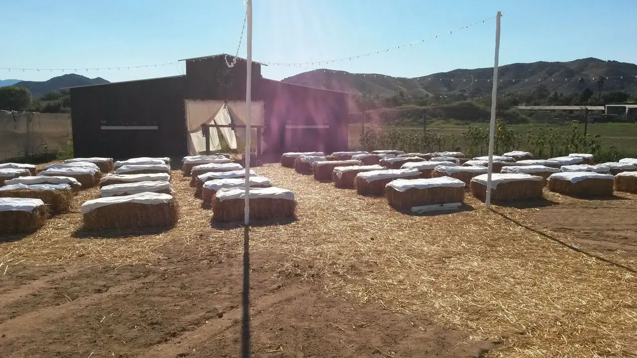 Country Hay Wedding Rows of hay bales covered with white cloth are arranged in front of a barn, with string lights overhead and mountains visible in the background.
