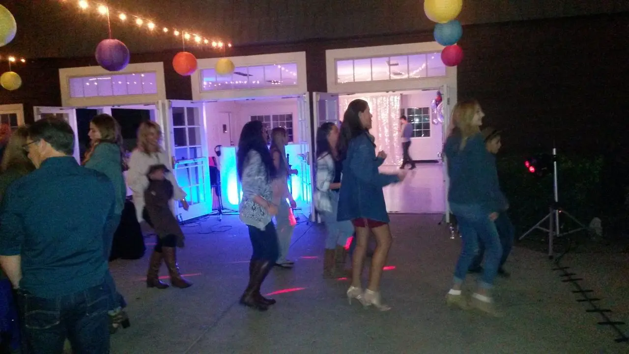 Karaoke Dj Kids Birthday Party A group of people dance together on a patio at night under string lights and colorful paper lanterns, with a building and open doors in the background.