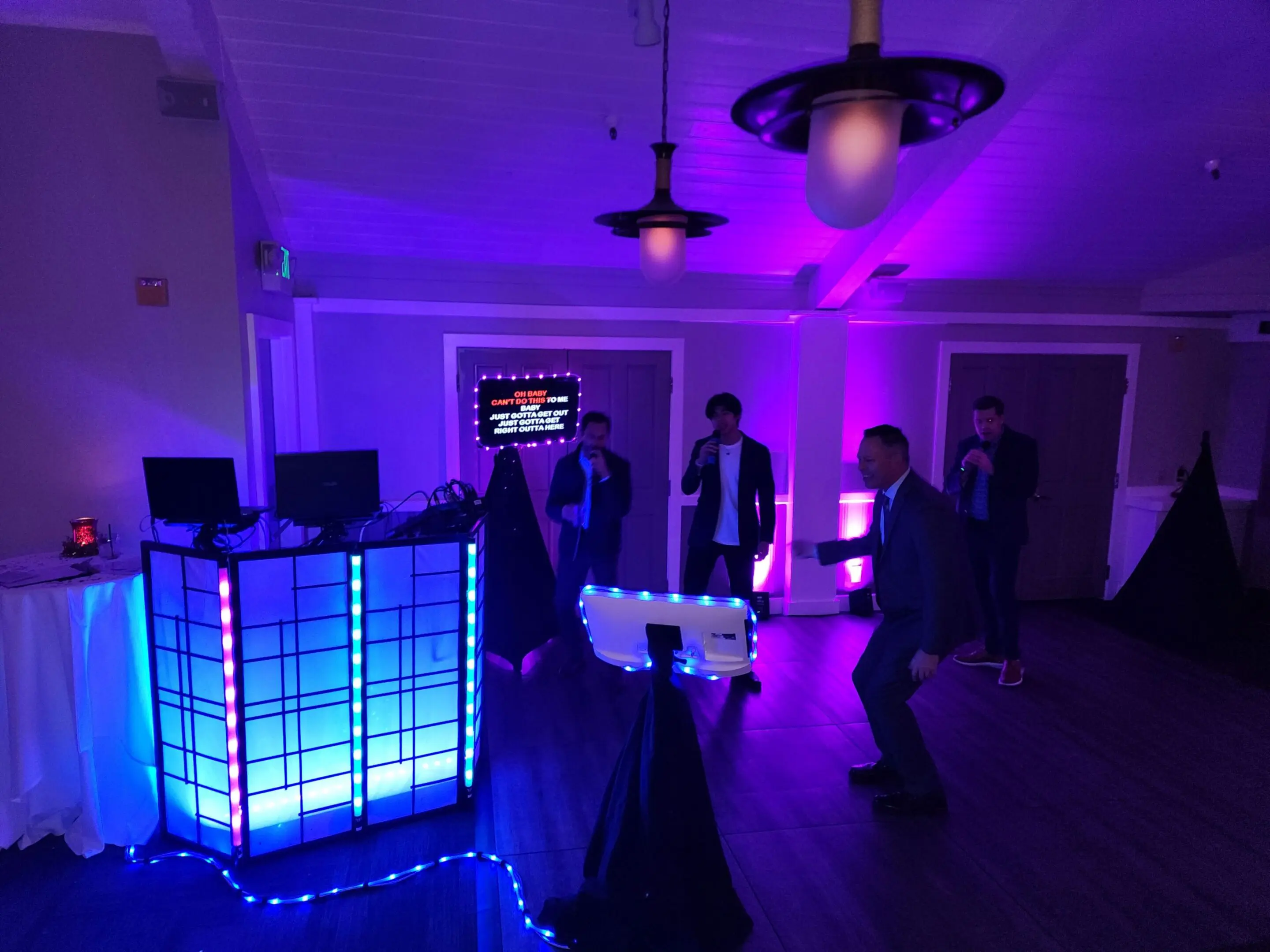 Our karaoke equipment set up with up lighting and guests singing karaoke songs near a karaoke DJ booth and a screen displaying lyrics.