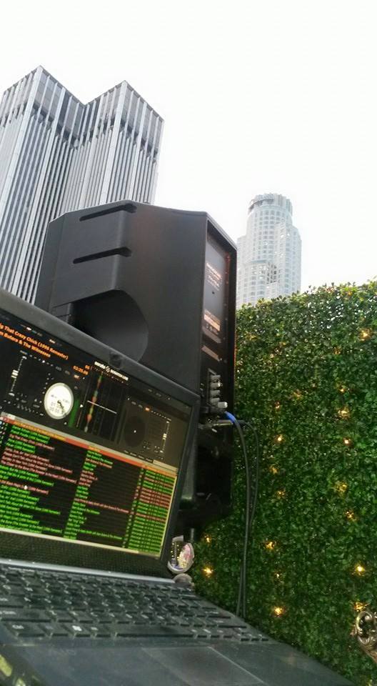 A DJ setup with a laptop and speaker in the foreground, with tall city buildings and a hedge wall in the background.