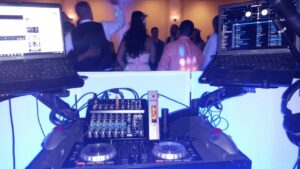 DJ setup with two laptops, a mixer, and turntables at a party with people dancing in the background.
