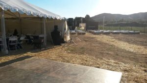 Outdoor event setup with a white tent containing chairs and tables on the left, a speaker and bales of hay on the right, and a wooden floor in the foreground, under a clear sky.