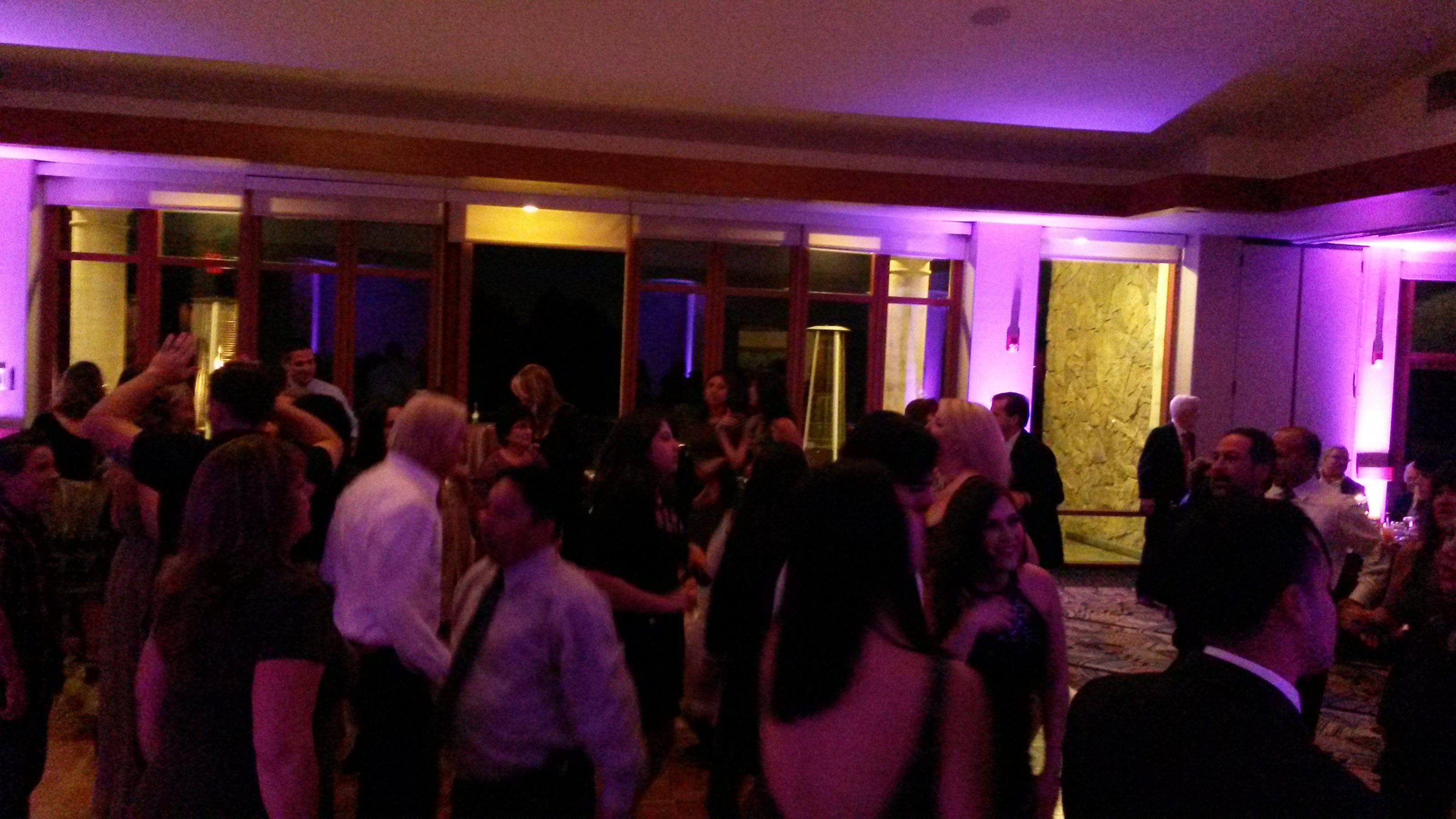 A group of people are dancing and socializing in a dimly lit room with purple lighting and large windows in the background.