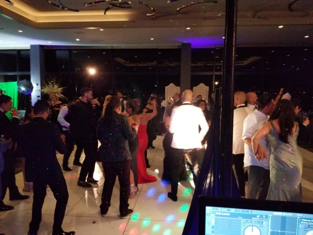 A group of people dressed in formal attire are dancing at an indoor event with dim lighting and colorful spotlights.