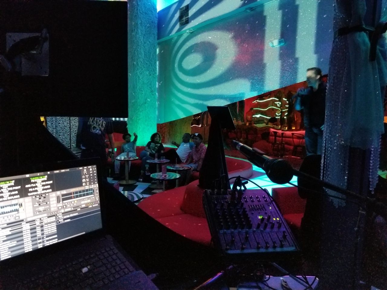 A DJ booth with a sound mixer and laptop overlooks a lounge where people are seated on red sofas while a person speaks or sings onstage under colorful lights.