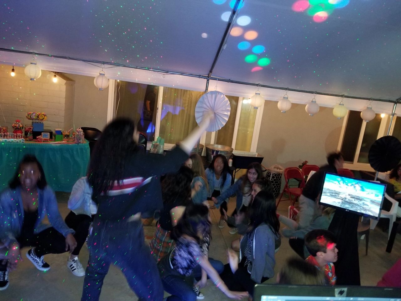 A group of people dance under colorful lights at an indoor party, with decorations and a table of snacks in the background.
