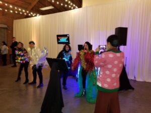 Four people perform karaoke in festive, shiny costumes while another person in a sweater films them with a phone in a room decorated with string lights and white curtains.