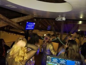 A group of people sing karaoke in a dimly lit room, facing a screen with song lyrics projected, under exposed wooden beams and a circular ceiling feature.
