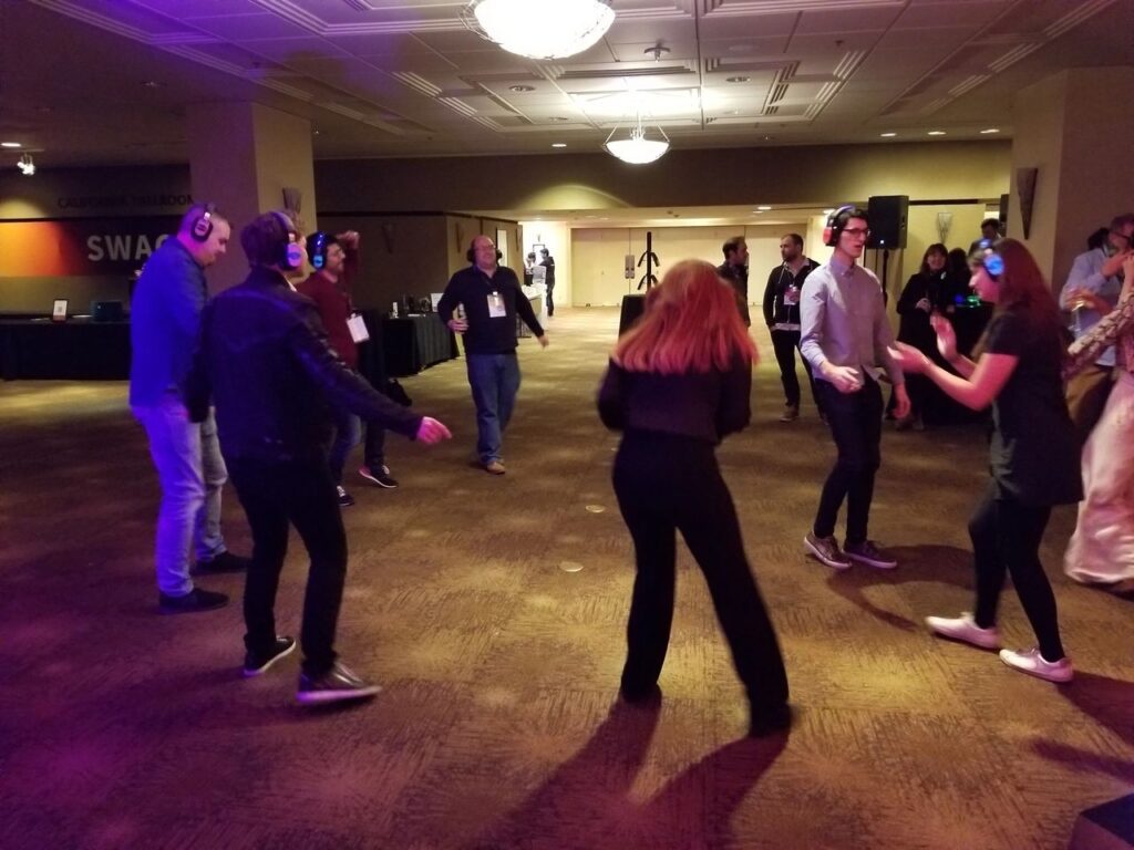 People wearing headphones dance in a dimly lit room, participating in a silent disco event. Other attendees stand or watch in the background.