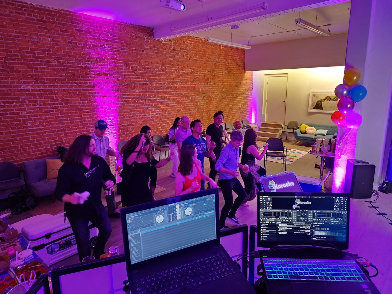 A group of people dance in a room with exposed brick walls and purple lighting, viewed from behind DJ equipment. Balloons and snacks are visible in the background.