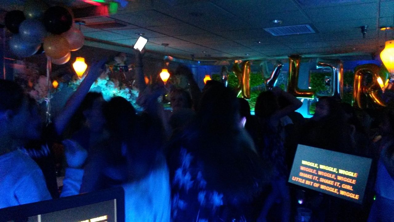 A group of people dance in a dimly lit room decorated with balloons and gold letter balloons, while karaoke lyrics are displayed on a screen in the foreground.