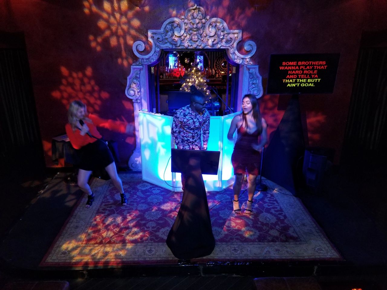 Three people are performing on a small stage with a decorative backdrop and colorful lights; lyrics are displayed on a screen to the right.