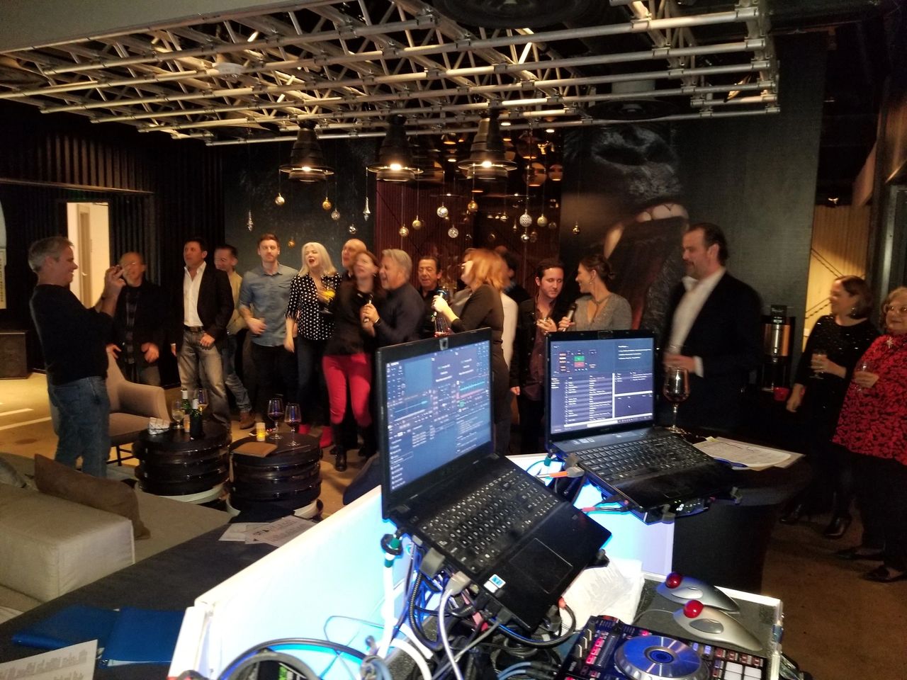 A group of people in semi-formal attire gather and socialize at an indoor event space, with DJ equipment and laptops set up in the foreground.