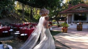 A bride in a white gown and veil holds a bouquet and smiles back while walking outdoors at a decorated wedding venue with round tables and red tablecloths.