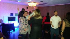 A group of people are dancing and socializing in a dimly lit room with a chandelier and DJ setup.