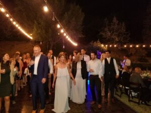 A group of people in formal attire walk together on a lit outdoor floor at night, with string lights overhead and trees in the background.