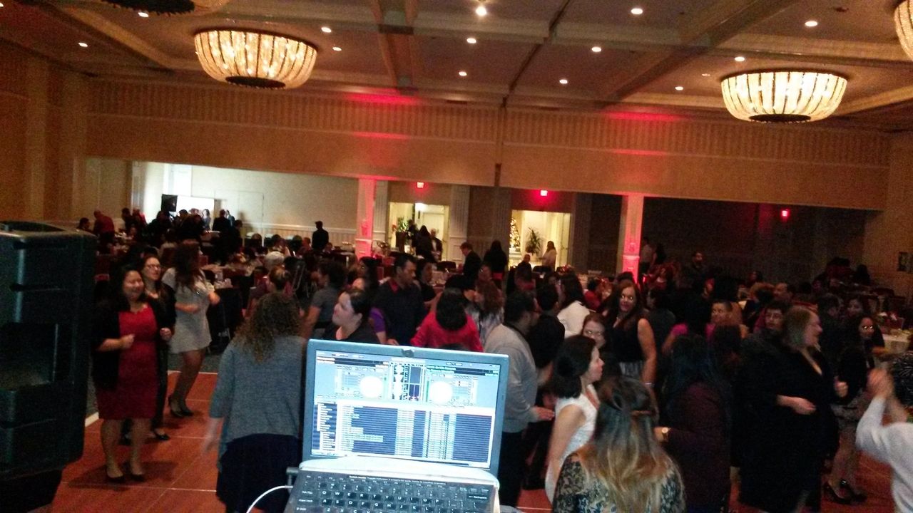 Large group of people dancing in a ballroom, seen from behind a DJ booth with a laptop displaying mixing software.