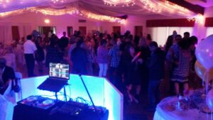 A large group of people are dancing in a decorated banquet hall with string lights, viewed from behind a DJ booth with turntables and a laptop.