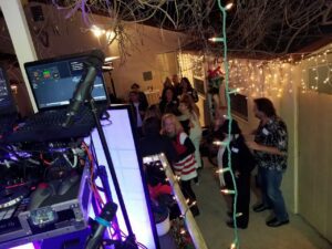 A group of people socialize and dance outdoors at night near a DJ booth, surrounded by string lights and festive decorations.