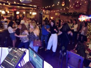 A group of people dance together in a warmly lit bar or restaurant, with music equipment visible in the foreground and festive decorations in the background.