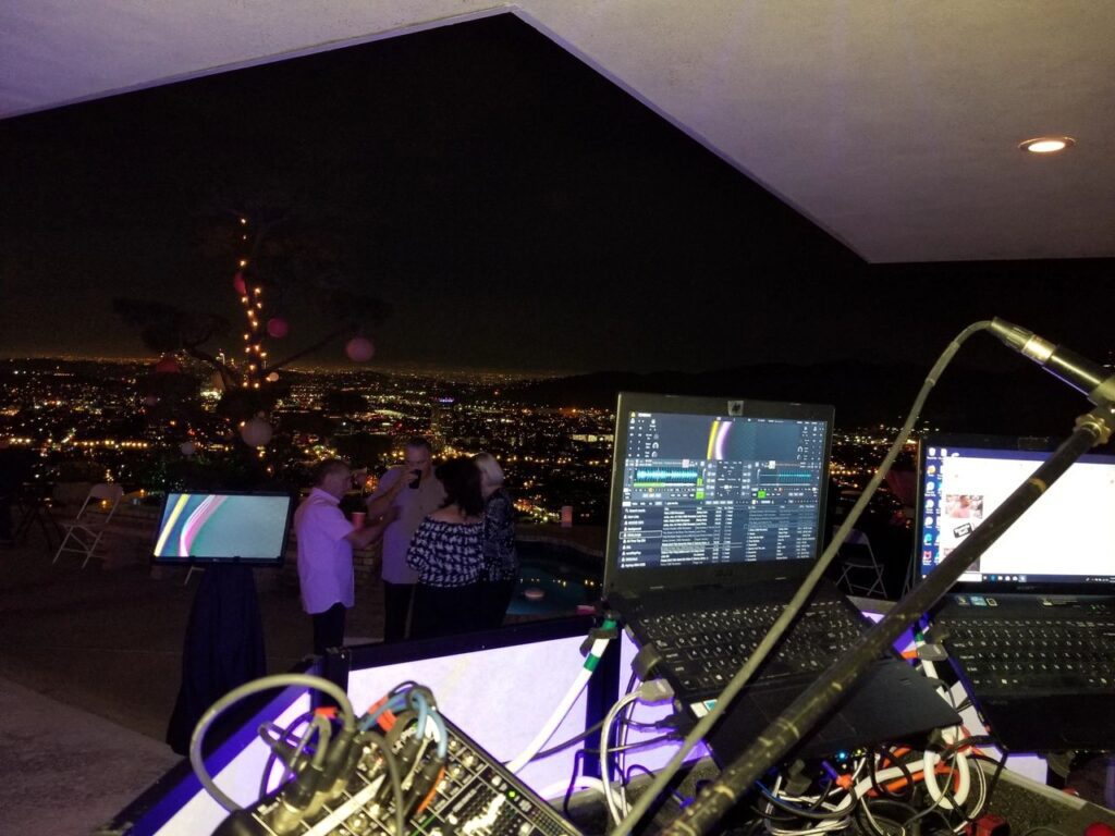 A nighttime rooftop scene shows DJ equipment in the foreground, with people dancing and city lights visible in the background.