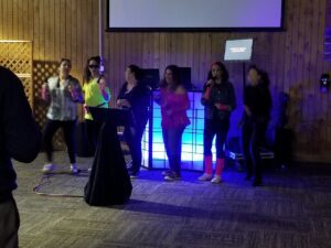 Moms in their 80s theme attire. singing karaoke, dancing on a stage in front of a lighted Party DJ booth and a karaoke screen in a wood-paneled room.