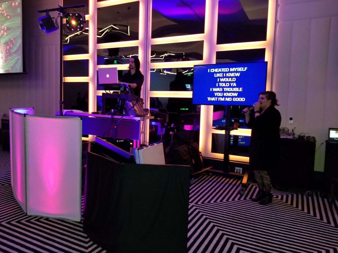 A woman sings karaoke while another operates a laptop at a DJ booth; song lyrics are displayed on a screen beside her in a room with colorful lighting.