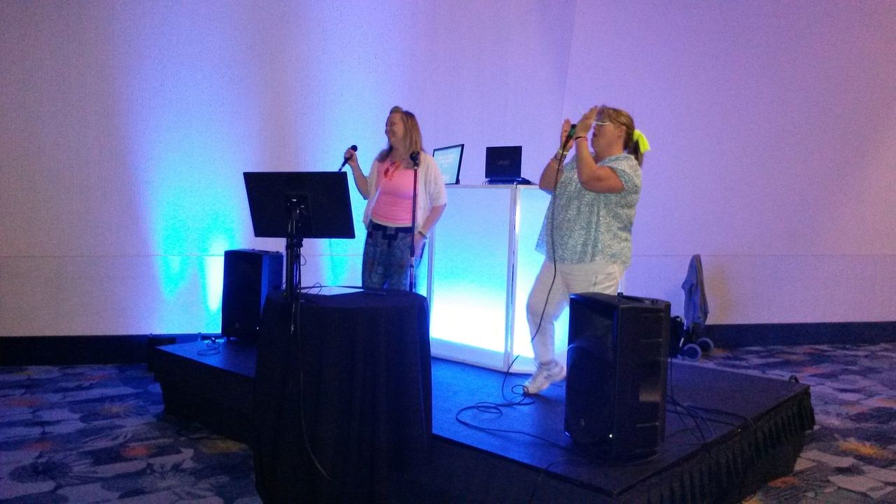 Two women stand on a small stage singing into microphones, with speakers and a screen in front of them and blue lighting behind them.