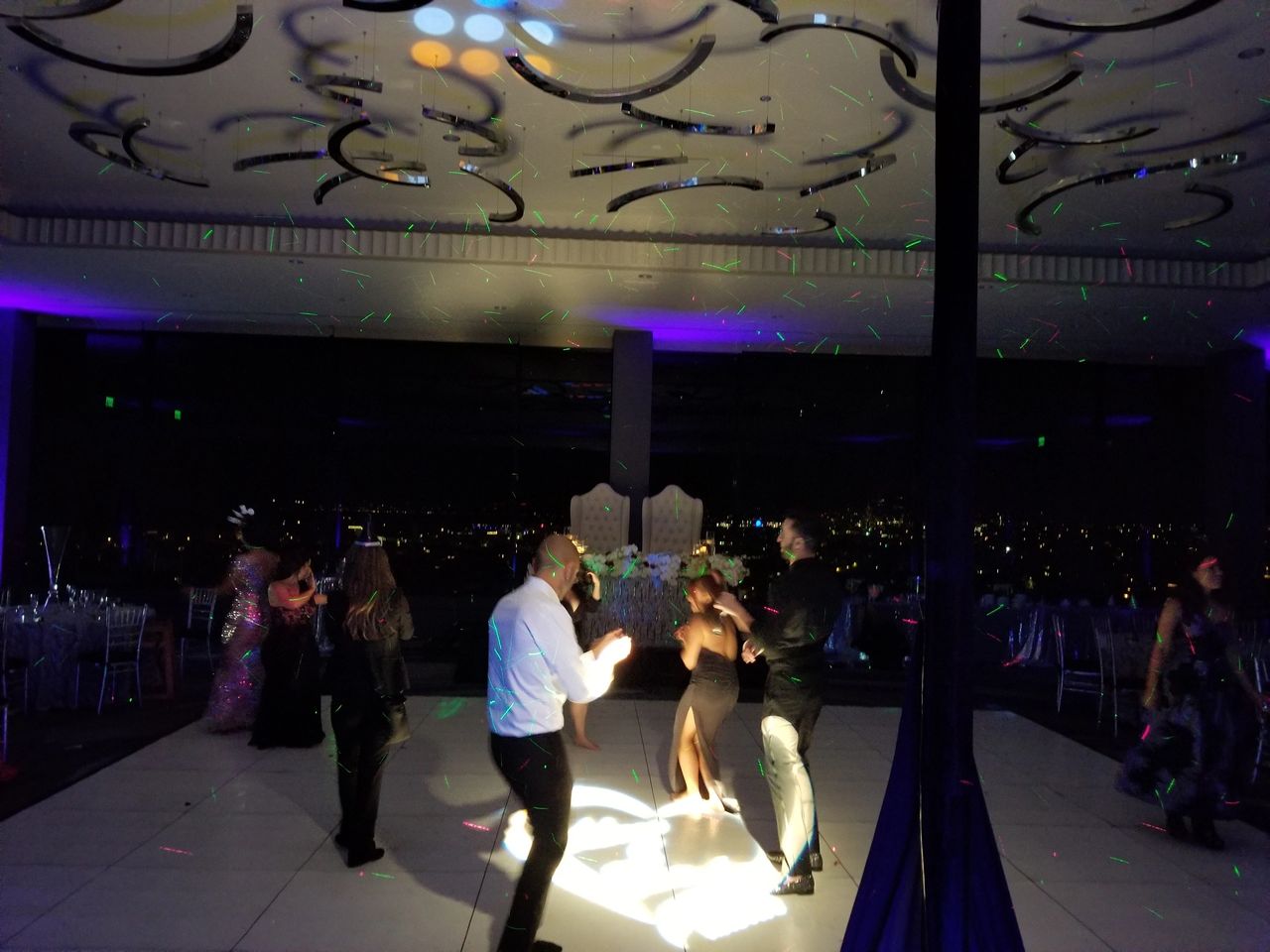 People are dancing on a dimly lit dance floor with colorful lights and cityscape visible through large windows in the background.