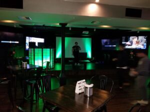 Dimly lit bar with empty tables, a central stage with green lighting, a person on stage, and multiple TV screens on the walls.