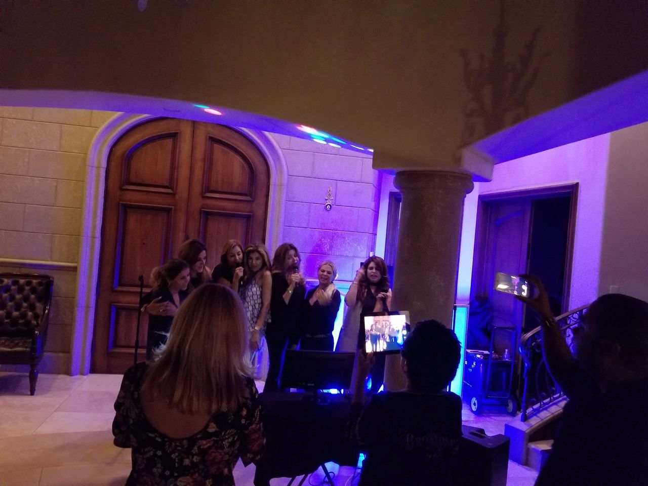 A group of women stand together singing or speaking into microphones in a warmly lit room, while others watch and take photos or videos.