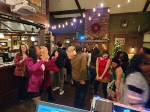 A group of people dance and socialize in a warmly lit indoor venue decorated with string lights and holiday ornaments.