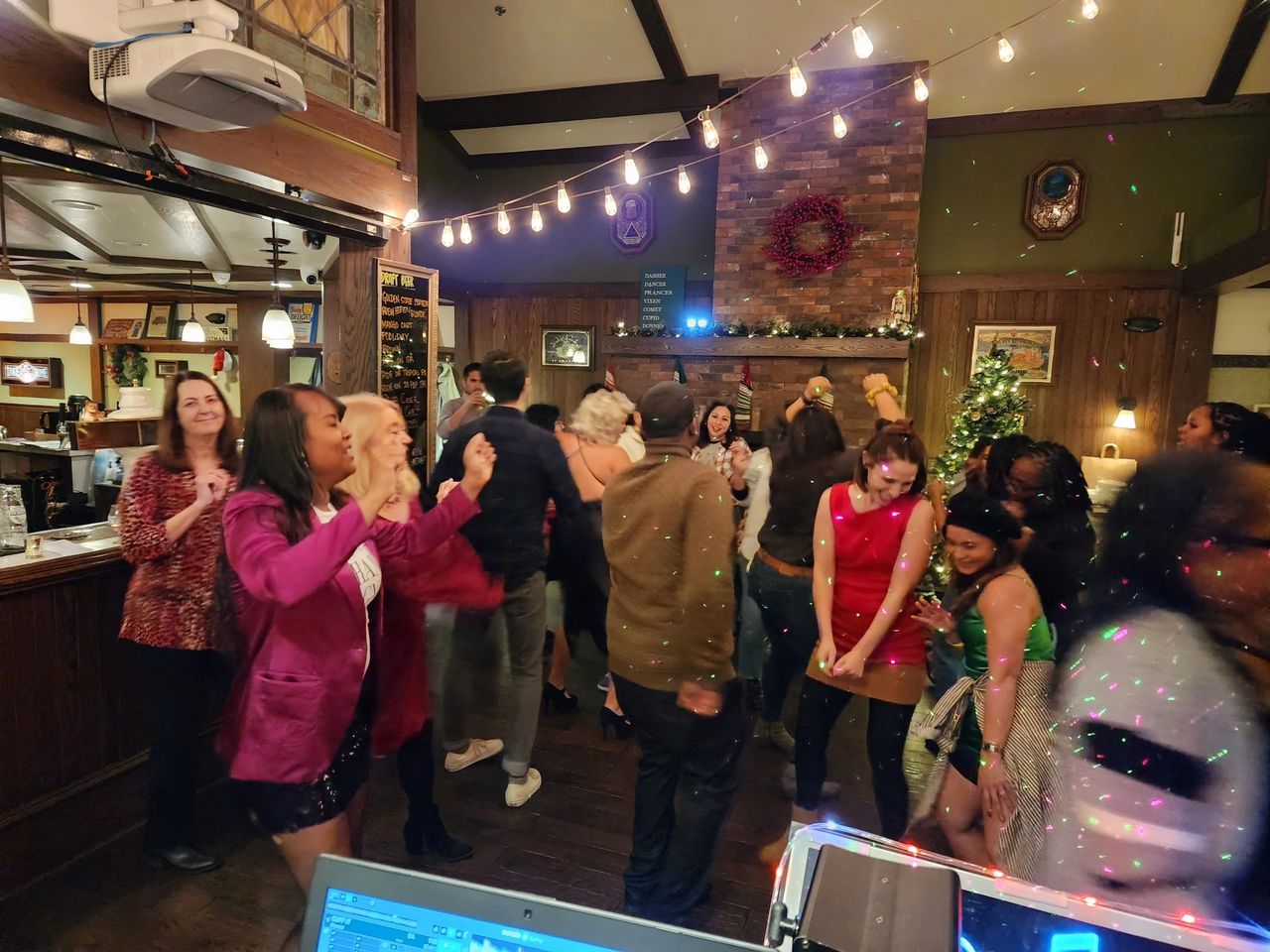 A group of people dance and socialize in a warmly lit indoor venue decorated with string lights and holiday ornaments.
