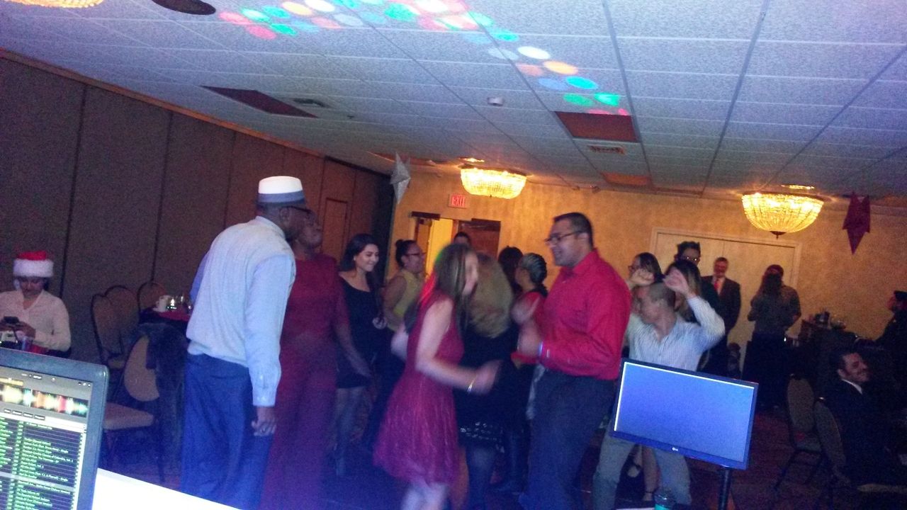 A group of people are dancing in a dimly lit event room with chandeliers, while others sit at tables. A DJ booth and computer screens are visible in the foreground.