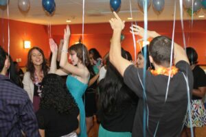 People are dancing and celebrating at an indoor party decorated with balloons and streamers.