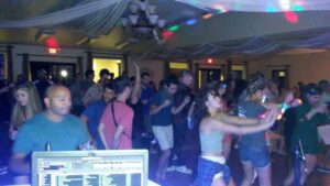 A crowd of people dance in a dimly lit indoor venue with colorful lights, as a DJ booth is visible in the foreground.