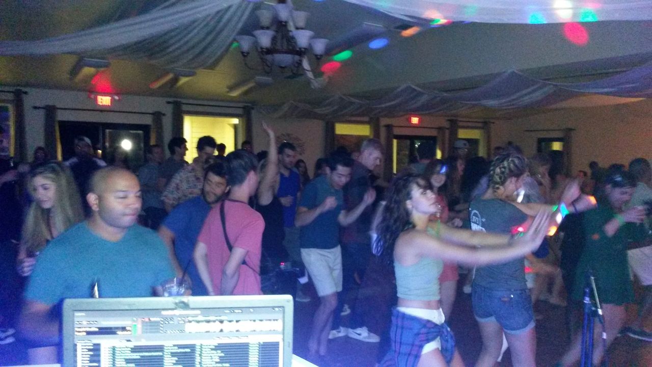 A crowd of people dance in a dimly lit indoor venue with colorful lights, as a DJ booth is visible in the foreground.