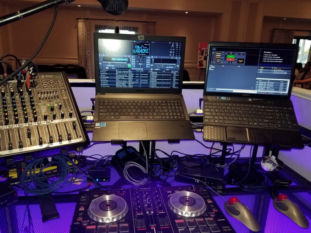 DJ and karaoke setup with a mixer, two laptops displaying DJ and karaoke software, a microphone, cables, and a DJ controller on a lit table.