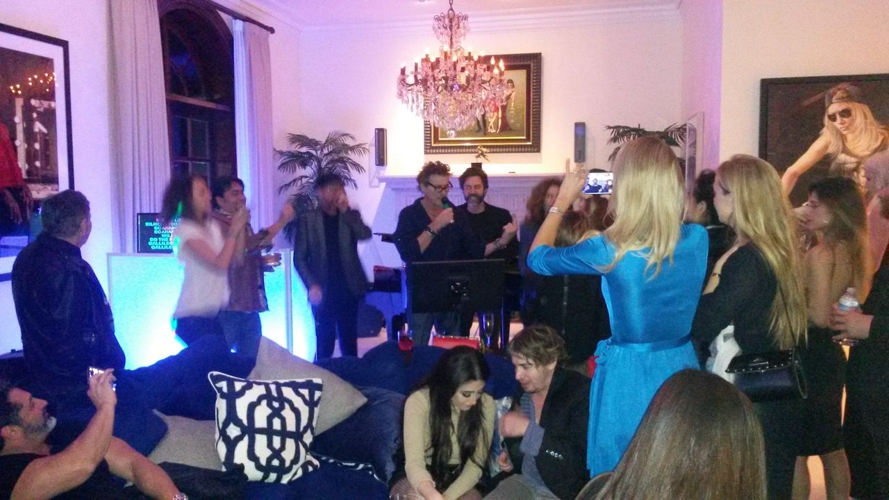 A group of people gathers in a living room, some standing and others sitting, with colorful lighting and a chandelier overhead. A woman in blue is taking a photo of the crowd.