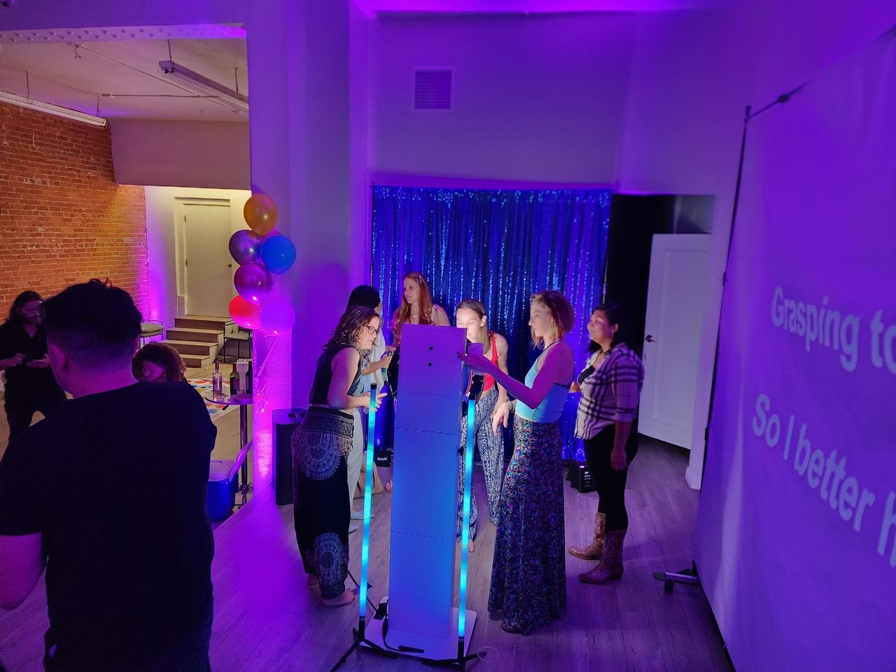 A group of people stand around a lighted photo booth at an indoor party with purple lighting; a karaoke screen is visible on the right.
