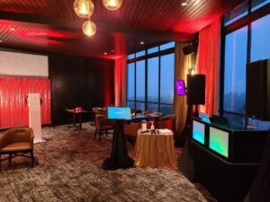 A modern event space with red lighting, tables and chairs, a DJ booth with colorful screens, a photo booth, and floor-to-ceiling windows overlooking a cityscape.